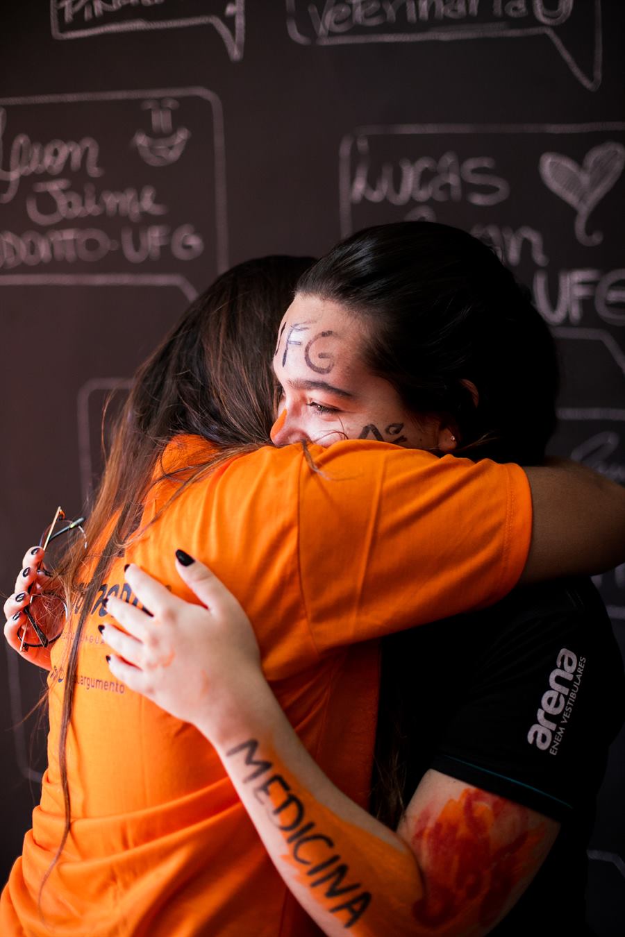 Duas alunas de camiseta laranja se abraçam em frente ao quadro de aprovados